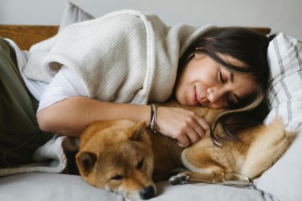 A woman peacefully napping on a sofa, embracing her Shiba Inu dog in a cozy indoor setting.